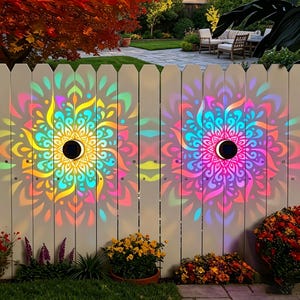 May include: Two circular solar-powered lights mounted on a white picket fence, projecting vibrant, multi-colored mandala patterns in shades of yellow, cyan, magenta, and purple onto the wooden slats, creating a glowing, symmetrical light display.