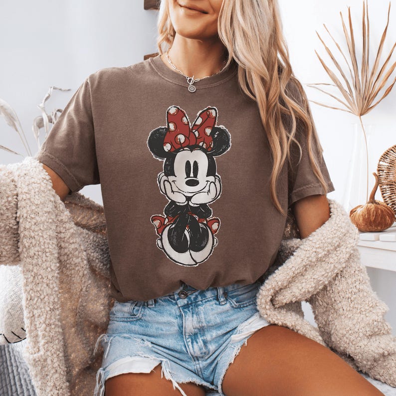 Pode incluir: Uma t-shirt castanha de manga curta com uma impress&atilde;o gr&aacute;fica desgastada da Minnie Mouse com um la&ccedil;o vermelho de bolinhas brancas. A personagem &eacute; representada com as m&atilde;os sob o queixo, vestindo um vestido vermelho com bolinhas brancas.