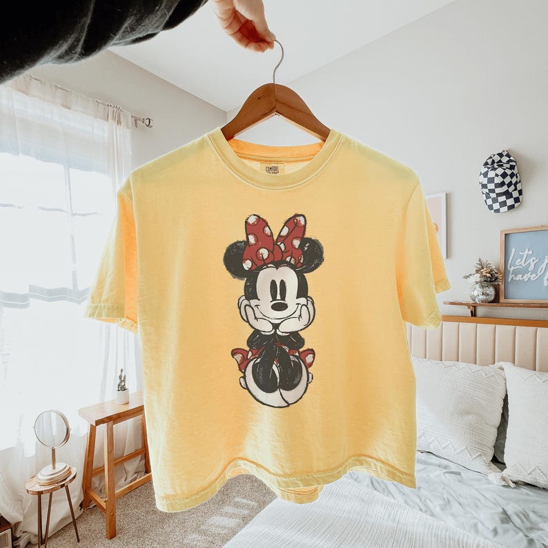 May include: A pale yellow short-sleeve t-shirt featuring a distressed black and white graphic of Minnie Mouse wearing a red and white polka dot bow. The character is depicted with hands resting under the chin, centered on the front of the shirt.
