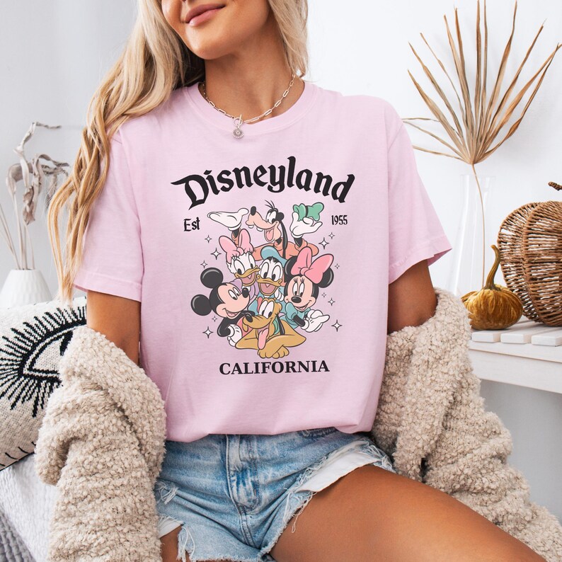 May include: A light pink short-sleeve t-shirt featuring a graphic print of Disney characters including Mickey Mouse, Minnie Mouse, Donald Duck, Daisy Duck, Goofy, and Pluto. The text reads Disneyland Est 1955 CALIFORNIA in black lettering.