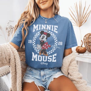 May include: A blue short-sleeve t-shirt featuring a graphic of Minnie Mouse inside a white and blue checkered circle. The text reads MINNIE MOUSE, 1928, and Disney. The design includes a red and white polka dot skirt and a small flower on her hat.
