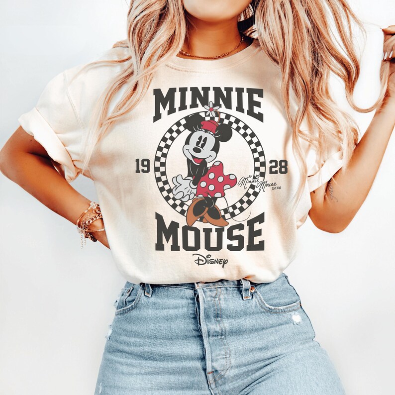 May include: A cream-colored t-shirt featuring a graphic of Minnie Mouse inside a black and white checkered circle. The text reads MINNIE MOUSE, 1928, and Disney. Minnie Mouse is wearing a red polka dot dress and a red bow with a small white flower.