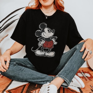 May include: A black short-sleeve t-shirt featuring a distressed graphic print of Mickey Mouse. The character is depicted in black, white, and red with a vintage-style texture. The shirt has a crew neckline and a relaxed fit.