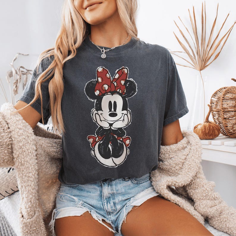 Pode incluir: Uma t-shirt de manga curta em cinzento-carv&atilde;o com uma impress&atilde;o gr&aacute;fica desgastada da Minnie Mouse com um la&ccedil;o vermelho de bolinhas brancas. A personagem &eacute; representada com as m&atilde;os sob o queixo, num estilo vintage com aspeto envelhecido.