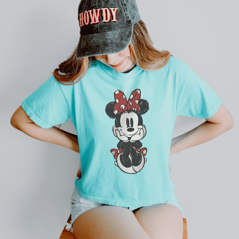 May include: A turquoise t-shirt featuring a graphic of Minnie Mouse with a red and white polka dot bow, paired with a distressed charcoal gray baseball cap with the word HOWDY embroidered in red and white letters on the front panel.
