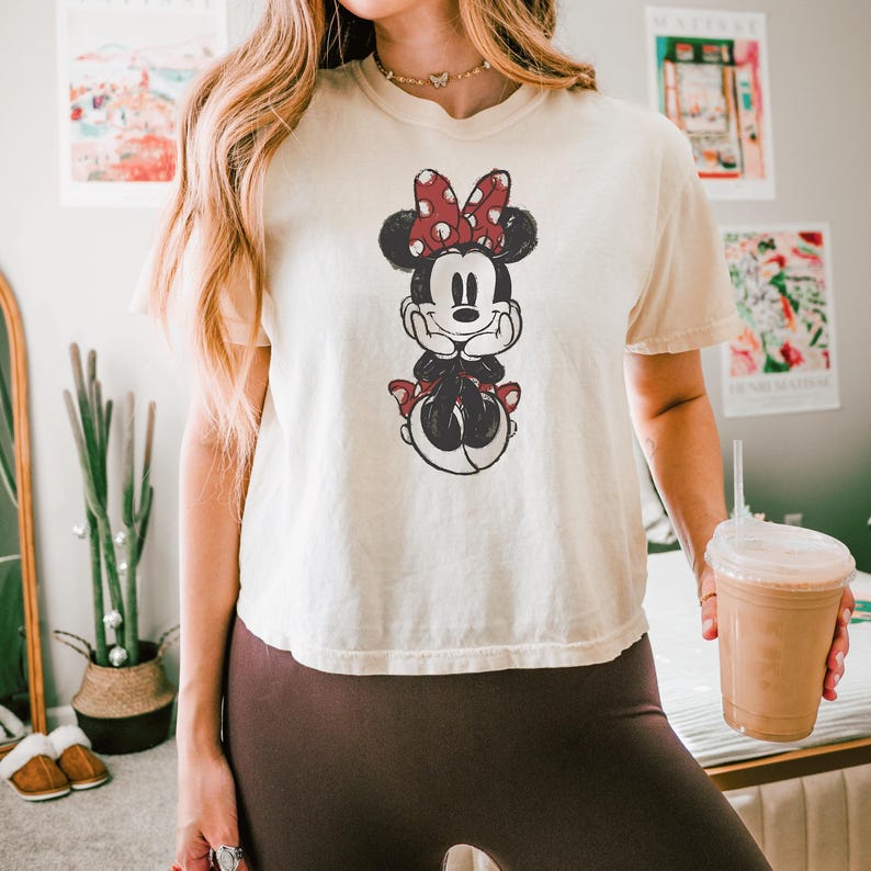 May include: A cream-colored short-sleeve t-shirt featuring a distressed black and red graphic print of Minnie Mouse. The character is depicted with her hands resting under her chin, wearing a red and white polka dot bow, centered on the chest.