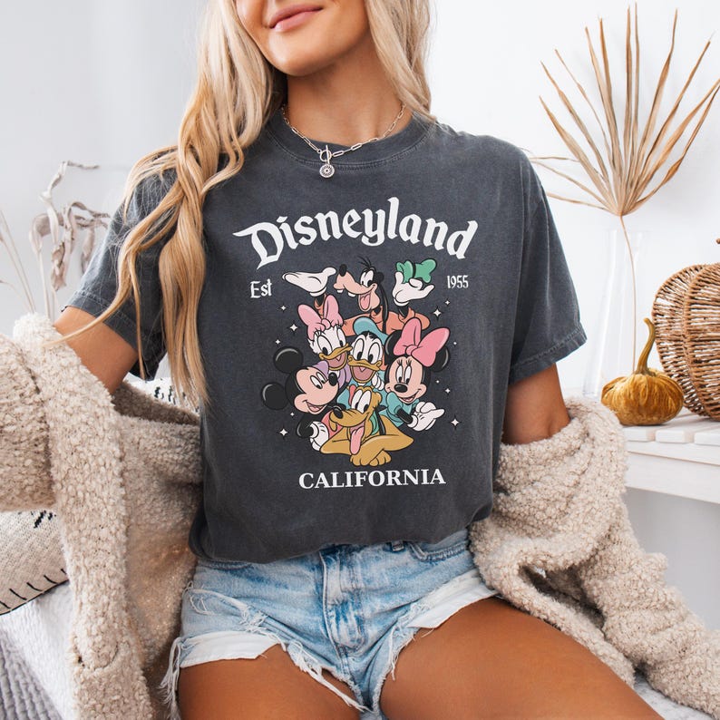 May include: A charcoal gray short-sleeve t-shirt featuring a graphic print of Mickey Mouse, Minnie Mouse, Donald Duck, Daisy Duck, Goofy, and Pluto. The text reads Disneyland Est 1955 California, surrounded by small white star accents.