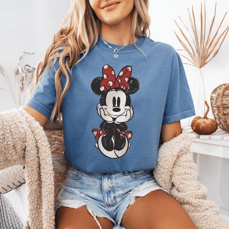 Pode incluir: Uma t-shirt azul de manga curta com uma impress&atilde;o gr&aacute;fica desgastada da Minnie Mouse. A personagem &eacute; representada com um la&ccedil;o vermelho de bolinhas brancas, orelhas pretas e um sorriso, com as m&atilde;os apoiadas sob o queixo.