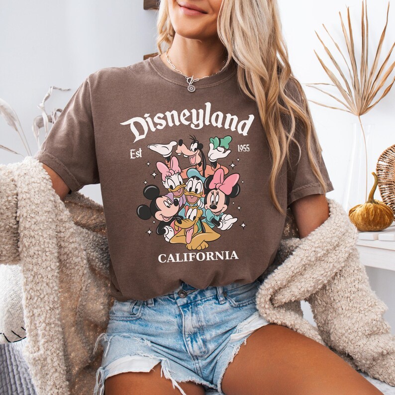 May include: A brown short-sleeve t-shirt featuring a graphic print of Disney characters including Mickey Mouse, Minnie Mouse, Donald Duck, Daisy Duck, Goofy, and Pluto. The text reads Disneyland Est 1955 CALIFORNIA with small star accents.
