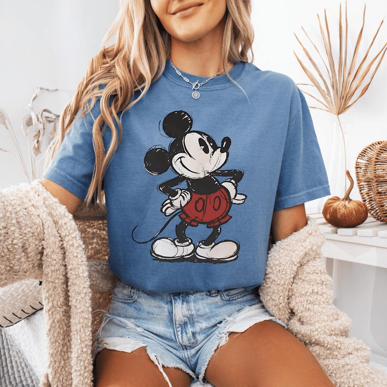 May include: A blue short-sleeved t-shirt featuring a vintage-style graphic of Mickey Mouse. The character is depicted in black, white, and red, wearing red shorts and white gloves. The shirt has a crew neckline and a relaxed, casual fit.