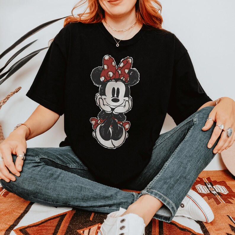 Pode incluir: Uma t-shirt preta de manga curta com uma estampa gr&aacute;fica da Minnie Mouse com um la&ccedil;o vermelho de bolinhas brancas. A personagem &eacute; retratada num estilo cl&aacute;ssico com as m&atilde;os sob o queixo, com uma textura desgastada de inspira&ccedil;&atilde;o vintage.