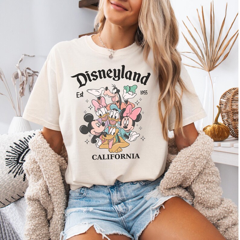 May include: A cream-colored short-sleeve t-shirt featuring a graphic print of Mickey Mouse, Minnie Mouse, Donald Duck, Daisy Duck, Goofy, and Pluto. The text reads Disneyland Est 1955 CALIFORNIA. The design includes small star accents.