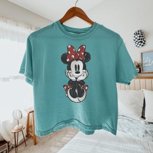 May include: A teal short-sleeve crew neck t-shirt featuring a centered graphic of Minnie Mouse. The character is depicted with her hands under her chin, wearing a red and white polka dot bow, and a matching red and white polka dot skirt.