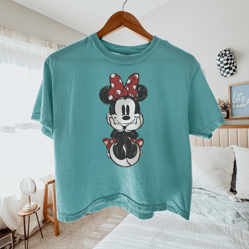 May include: A teal short-sleeve crew neck t-shirt featuring a centered graphic of Minnie Mouse. The character is depicted with her hands under her chin, wearing a red and white polka dot bow, and a matching red and white polka dot skirt.