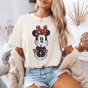 Pode incluir: Uma t-shirt de manga curta cor de creme com uma impress&atilde;o gr&aacute;fica desgastada da Minnie Mouse. A personagem &eacute; representada a preto e branco com um la&ccedil;o vermelho de bolinhas brancas e sapatos vermelhos a condizer, com o queixo nas m&atilde;os.