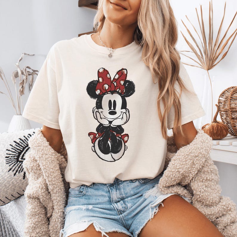 Pode incluir: Uma t-shirt de manga curta cor de creme com uma impress&atilde;o gr&aacute;fica desgastada da Minnie Mouse. A personagem &eacute; representada a preto e branco com um la&ccedil;o vermelho de bolinhas brancas e sapatos vermelhos a condizer, com o queixo nas m&atilde;os.