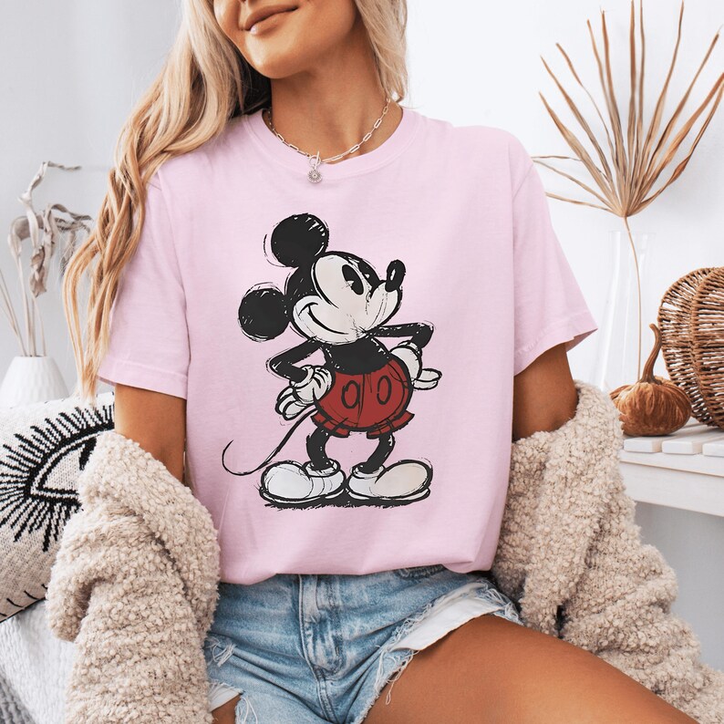 May include: A light pink short-sleeved t-shirt featuring a black and white graphic illustration of Mickey Mouse wearing red shorts. The character is depicted in a classic style with hands on hips, centered on the front of the garment.
