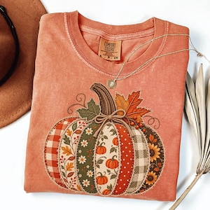 May include: A terracotta-colored t-shirt featuring a large pumpkin graphic composed of various patterned fabric segments including plaid, floral, and polka dot prints. The pumpkin is accented with a twine bow, leaves, and berries.
