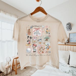 K&ouml;nnte beinhalten: Ein beiges T-Shirt mit einem Grafikdruck von Walt Disney World-Symbolen, darunter Mickey Maus, Minnie Maus, Donald Duck, Daisy Duck, Goofy, Chip und Chap, das Schloss und Spaceship Earth, mit dem Text The Most Magical Place on Earth 2026.
