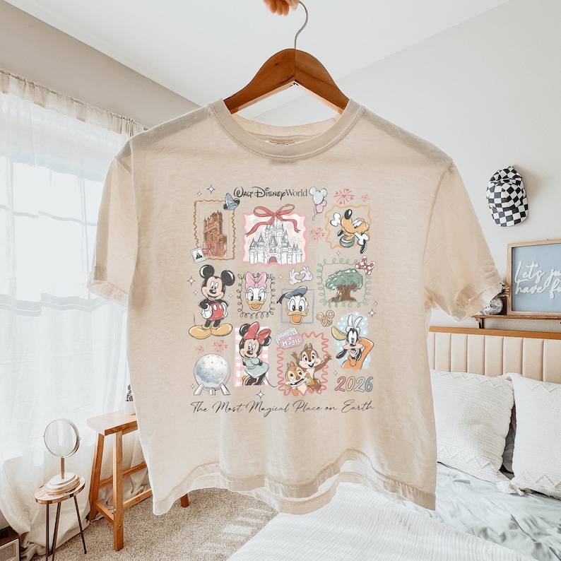 K&ouml;nnte beinhalten: Ein beiges T-Shirt mit einem Grafikdruck von Walt Disney World-Symbolen, darunter Mickey Maus, Minnie Maus, Donald Duck, Daisy Duck, Goofy, Chip und Chap, das Schloss und Spaceship Earth, mit dem Text The Most Magical Place on Earth 2026.