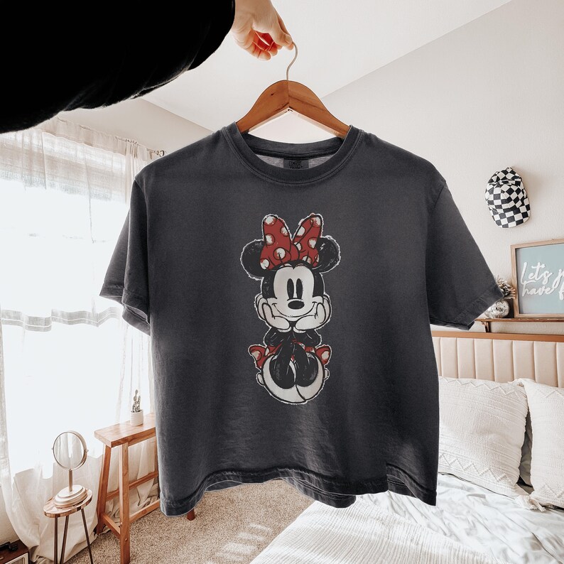 May include: A charcoal gray short-sleeve t-shirt featuring a distressed graphic print of Minnie Mouse on the front. The character is depicted with her signature red and white polka dot bow, resting her chin on her hands in a centered position.