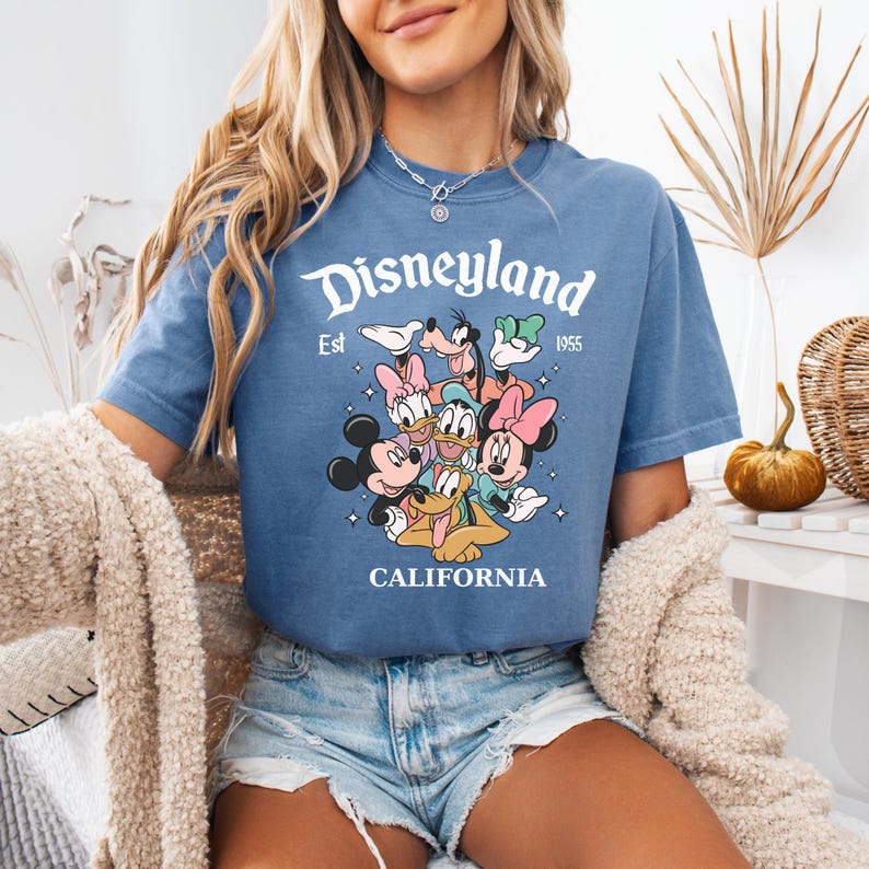May include: A blue short-sleeve t-shirt featuring a graphic print of Disney characters including Mickey Mouse, Minnie Mouse, Donald Duck, Daisy Duck, Goofy, and Pluto. The text reads Disneyland Est 1955 CALIFORNIA in white lettering.