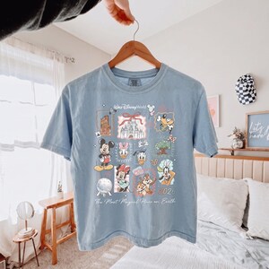 K&ouml;nnte beinhalten: Ein hellblaues T-Shirt mit einem Grafikdruck von Walt Disney World-Symbolen, darunter Mickey Maus, Minnie Maus, Donald Duck, Daisy Duck, Goofy, Chip und Chap, das Cinderella-Schloss und Spaceship Earth, mit dem Text 2026 und The Most Magical Place on Earth.