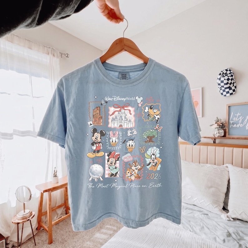 K&ouml;nnte beinhalten: Ein hellblaues T-Shirt mit einem Grafikdruck von Walt Disney World-Symbolen, darunter Mickey Maus, Minnie Maus, Donald Duck, Daisy Duck, Goofy, Chip und Chap, das Cinderella-Schloss und Spaceship Earth, mit dem Text 2026 und The Most Magical Place on Earth.