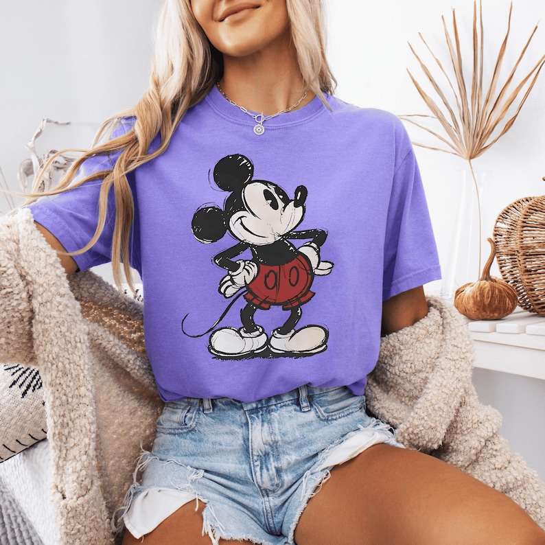 May include: A purple short-sleeved t-shirt featuring a vintage-style graphic of Mickey Mouse. The character is depicted in black, white, and red with a distressed, textured print. The shirt has a crew neckline and a relaxed, casual fit.