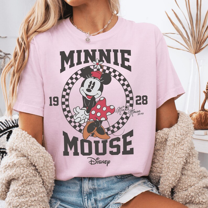 May include: A light pink t-shirt featuring a graphic of Minnie Mouse inside a black and white checkered circle. The text reads MINNIE MOUSE, 1928, and Disney. Minnie Mouse wears a red hat and a red polka dot dress. The design includes the signature Minnie Mouse XOXO.
