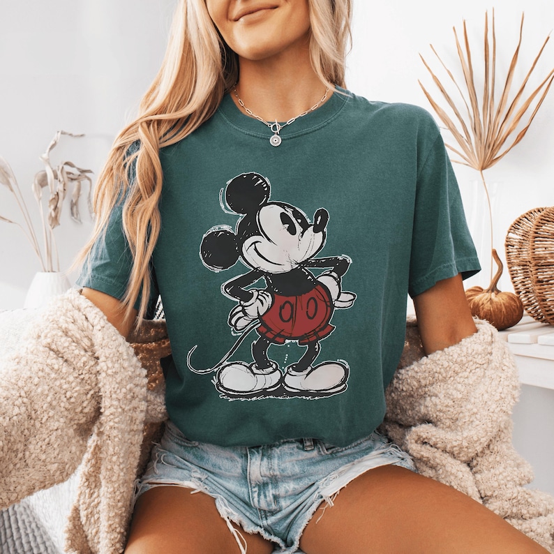 May include: A dark teal short-sleeve t-shirt featuring a distressed graphic print of Mickey Mouse on the front. The character is depicted in a classic pose with black ears, a white face, and red shorts. The shirt has a crew neckline and a relaxed fit.