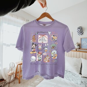 K&ouml;nnte beinhalten: Ein hellviolettes T-Shirt mit einem Grafikdruck von Walt Disney World-Symbolen und Charakteren wie Mickey Maus, Minnie Maus, Donald Duck, Daisy Duck, Goofy, Chip und Chap, dem Schloss und dem Text The Most Magical Place on Earth 2026.