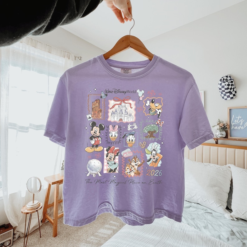 K&ouml;nnte beinhalten: Ein hellviolettes T-Shirt mit einem Grafikdruck von Walt Disney World-Symbolen und Charakteren wie Mickey Maus, Minnie Maus, Donald Duck, Daisy Duck, Goofy, Chip und Chap, dem Schloss und dem Text The Most Magical Place on Earth 2026.
