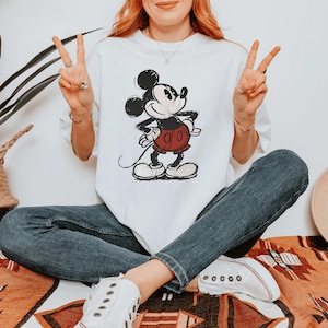 May include: A white short-sleeve t-shirt featuring a vintage-style graphic of Mickey Mouse in black and white with red shorts. The character is depicted in a classic pose with hands on hips. The shirt is paired with dark blue denim jeans and white sneakers.