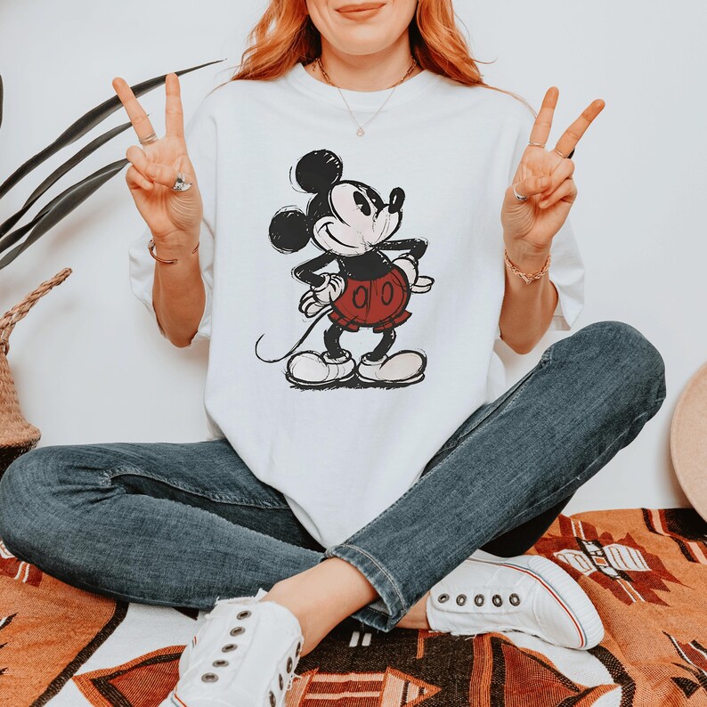 May include: A white short-sleeve t-shirt featuring a vintage-style graphic of Mickey Mouse in black and white with red shorts. The character is depicted in a classic pose with hands on hips. The shirt is paired with dark blue denim jeans and white sneakers.