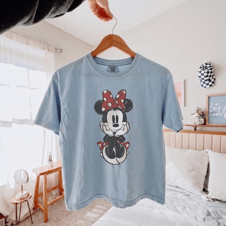 May include: A light blue short-sleeve t-shirt featuring a centered graphic of Minnie Mouse with her hands resting under her chin. The character wears a red bow with white polka dots and a matching red skirt. The shirt has a crew neckline.