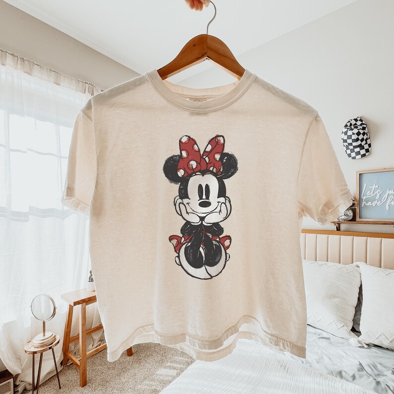 May include: A cream-colored short-sleeve t-shirt featuring a distressed graphic print of Minnie Mouse. The character is depicted with her hands under her chin, wearing her signature red and white polka dot bow, rendered in black, white, and red ink.