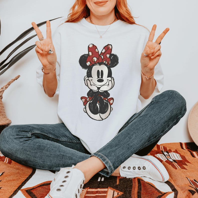 Pode incluir: Uma t-shirt branca de manga curta com uma estampa gr&aacute;fica desgastada de um rato de desenho animado com um la&ccedil;o vermelho de bolinhas brancas, combinada com cal&ccedil;as de ganga azul-escuras e t&eacute;nis de lona brancos com atacadores e sola de borracha.