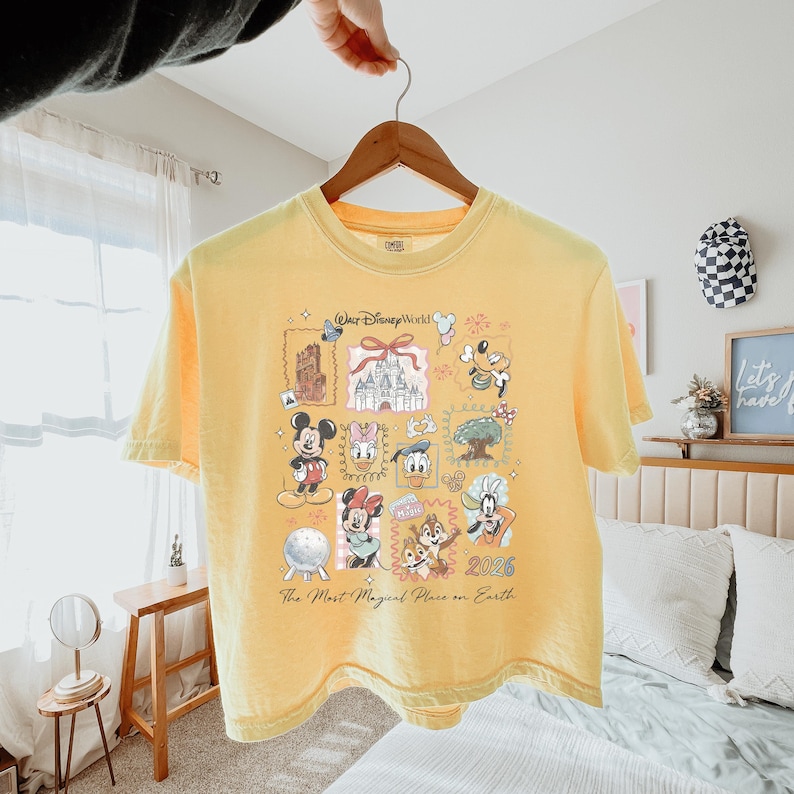 K&ouml;nnte beinhalten: Ein blassgelbes T-Shirt mit einem Grafikdruck von Walt Disney World-Symbolen und Charakteren wie Mickey Maus, Minnie Maus, Donald Duck, Daisy Duck, Goofy sowie Chip und Chap, inklusive dem Text "The Most Magical Place on Earth" und "2026".