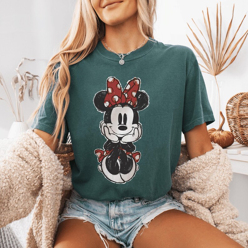 Pode incluir: Uma t-shirt de manga curta em verde-azulado escuro com uma estampa gr&aacute;fica desgastada da Minnie Mouse. A personagem &eacute; retratada com o seu la&ccedil;o vermelho de bolinhas brancas, orelhas pretas e luvas brancas, apoiando o queixo nas m&atilde;os.