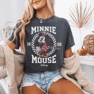 May include: A charcoal gray short-sleeve t-shirt featuring a graphic of Minnie Mouse inside a black and white checkered circle. The text reads MINNIE MOUSE, 19, 28, and Disney. The design includes a red polka dot skirt and yellow shoes.