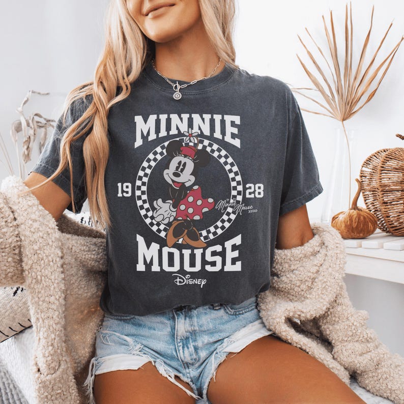 May include: A charcoal gray short-sleeve t-shirt featuring a graphic of Minnie Mouse inside a black and white checkered circle. The text reads MINNIE MOUSE, 19, 28, and Disney. The design includes a red polka dot skirt and yellow shoes.