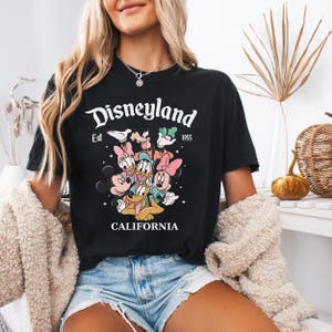 May include: A black short-sleeved t-shirt featuring a graphic print of Disney characters including Mickey Mouse, Minnie Mouse, Donald Duck, Daisy Duck, Goofy, and Pluto. The text reads Disneyland Est 1955 CALIFORNIA with small star accents.