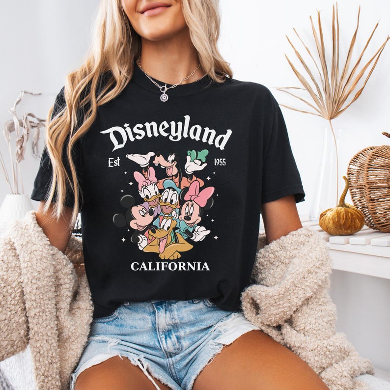 May include: A black short-sleeved t-shirt featuring a graphic print of Disney characters including Mickey Mouse, Minnie Mouse, Donald Duck, Daisy Duck, Goofy, and Pluto. The text reads Disneyland Est 1955 CALIFORNIA with small star accents.