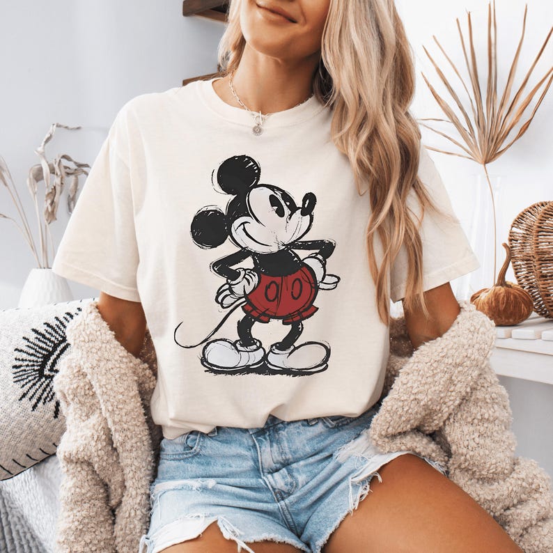 May include: A cream-colored short-sleeve t-shirt featuring a black and red graphic illustration of Mickey Mouse. The character is depicted in a classic pose with hands on hips, wearing red shorts and large shoes, rendered in a sketch-like style.