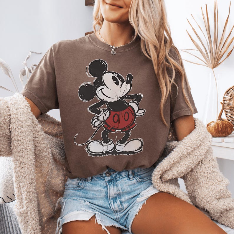 May include: A brown short-sleeved t-shirt featuring a distressed graphic print of Mickey Mouse in black, white, and red. The character is depicted standing with hands on hips, wearing red shorts and white shoes, centered on the front of the shirt.