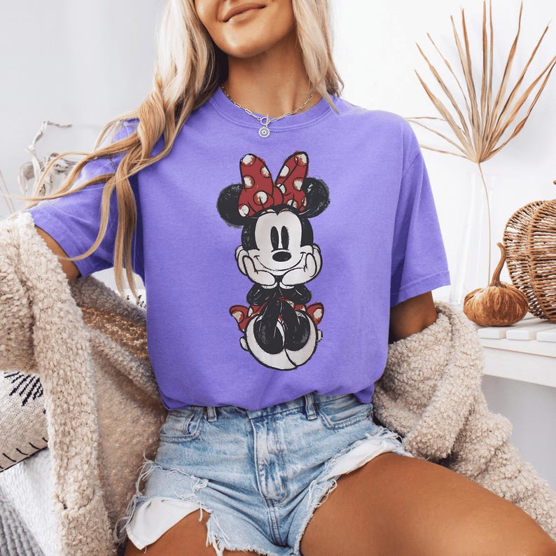 Pode incluir: Uma t-shirt roxa de manga curta com uma impress&atilde;o gr&aacute;fica desgastada da Minnie Mouse. A personagem &eacute; representada com um la&ccedil;o vermelho de bolinhas brancas, orelhas pretas e luvas brancas, com o queixo apoiado nas m&atilde;os ao centro.