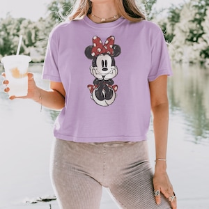 May include: A light purple short-sleeved t-shirt featuring a graphic print of a cartoon mouse with a red and white polka dot bow, resting its chin on its hands. The shirt has a crew neckline and a relaxed fit, showcasing a distressed ink design.