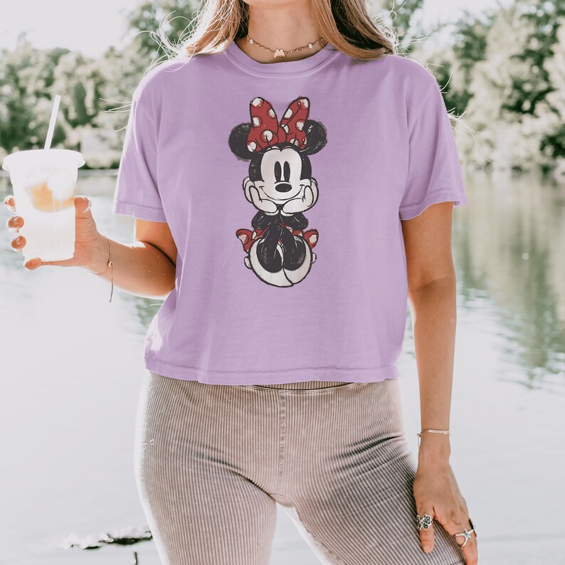 May include: A light purple short-sleeved t-shirt featuring a graphic print of a cartoon mouse with a red and white polka dot bow, resting its chin on its hands. The shirt has a crew neckline and a relaxed fit, showcasing a distressed ink design.