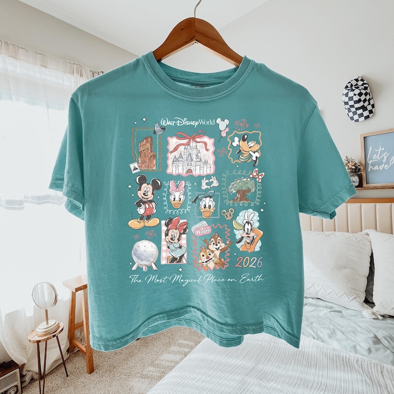 K&ouml;nnte beinhalten: Ein blaugr&uuml;nes Kurzarm-T-Shirt mit einem Grafikdruck von Walt Disney World-Figuren, dem Schloss und Wahrzeichen. Der Text lautet Walt Disney World, 2026 und The Most Magical Place on Earth. Das Design zeigt Mickey, Minnie, Donald, Daisy, Goofy sowie Chip und Chap.